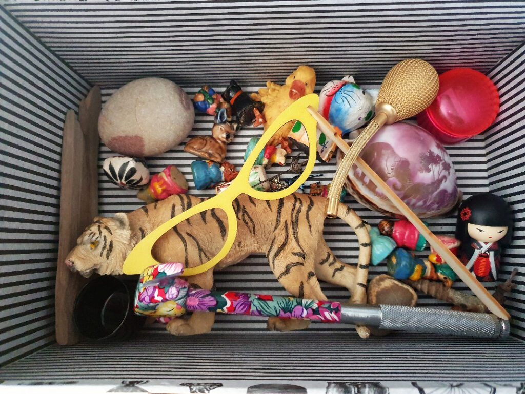 A box containing objects, including a plastic toy tiger, a pair of yellow toy glasses, a hammer with a floral pattern, ceramic toy figures, a shell with African wildlife embossed.